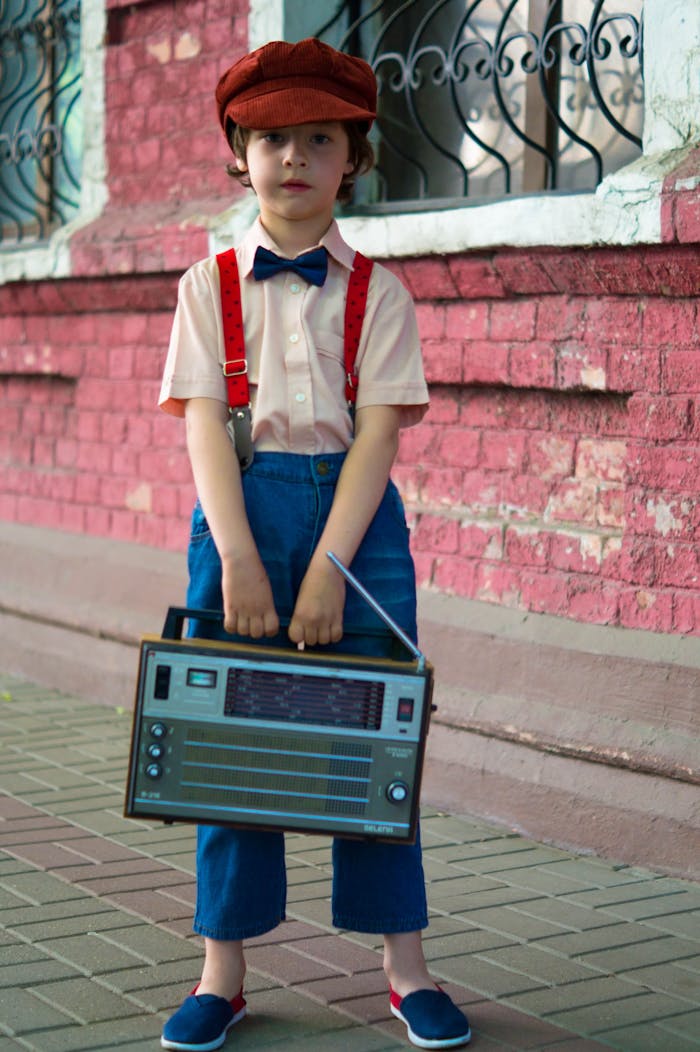 Photo of Boy Holding Radio and Standing Beside of Red House